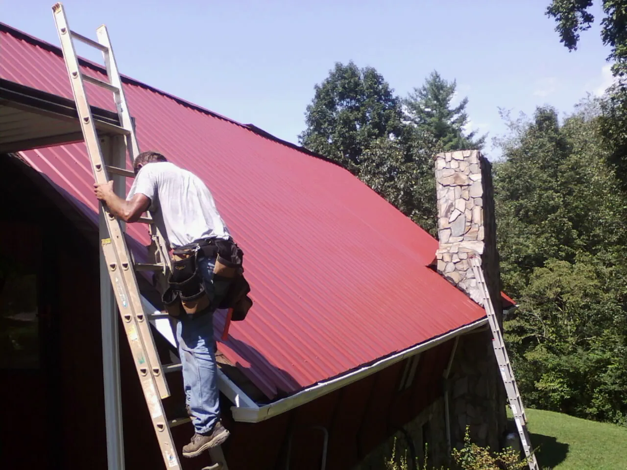 Roofer on a ladder beside a red metal roof and stone chimney — Soffit & Fascia Repair by Roofing Brothers in Far Hills